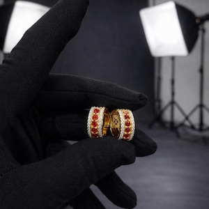May include: Two gold-colored hoop earrings with red gemstones and white diamonds. The earrings are held by a gloved hand. The background is dark with studio lighting.