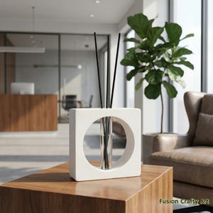 May include: A white, square-shaped diffuser with a circular opening, holding a glass vial and black reeds. The diffuser sits on a wooden table. A large green plant and a leather armchair are in the background.