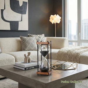 May include: An hourglass with a wooden frame and black sand sits on a concrete coffee table. The hourglass is framed by black metal rods. The table also holds books, decorative objects, and a gold-toned tray. The background includes a cream-colored sofa and a large abstract painting.
