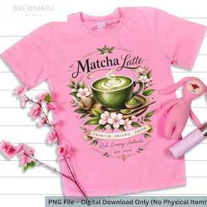May include: A pink t-shirt featuring a Matcha Latte design. The graphic includes a green cup of matcha, flowers, and the text "Matcha Latte". The design also includes the text "Premium Organic Fresh" and "Rich Creamy Authentic".