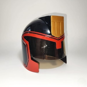 Könnte beinhalten: Ein schwarzer und roter Helm mit goldenem Akzent oben. Der Helm hat ein klares Visier und rote Zierleisten. Das Design ist elegant und futuristisch, mit glänzendem Finish. Der Helm ist wahrscheinlich für Cosplay oder Kostüme gedacht.