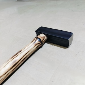May include: A black metal sledgehammer with a rectangular head and a wooden handle. The handle has a light brown color with dark brown wood grain patterns. The sledgehammer is designed for heavy-duty tasks and construction work.
