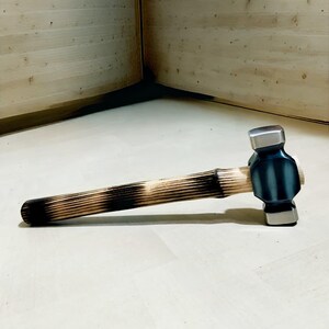 May include: A hammer with a light-coloured wooden handle featuring a dark, burnt design. The hammerhead is a dark blue-grey colour with a silver striking face. The tool is designed for striking and shaping materials.