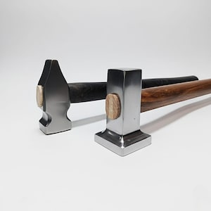 May include: Two metal hammers with wooden handles. One hammer has a dark handle, the other a light brown handle. The hammer heads are silver and have a wooden accent. The hammers are on a white background.