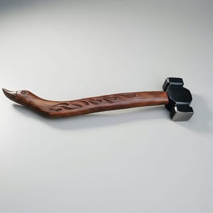 May include: A unique hammer with a dark wood handle carved in the shape of a bird's head and neck. The handle features carved flame-like designs. The hammerhead is black and rectangular, made of metal.