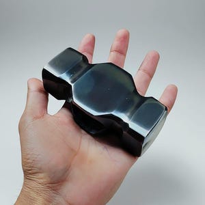 May include: A black, polished hammer with a rectangular head and a rounded handle. The hammer is held in a person's hand, showcasing its size and shape. The tool has a reflective surface, suggesting it is made of metal.