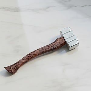 Può includere: Un martello artigianale con una testa in argento lucido e un manico in legno intagliato. Il manico presenta un'intricata lavorazione a nodi celtici e un motivo a raggiera alla base. Il martello è un oggetto decorativo unico.