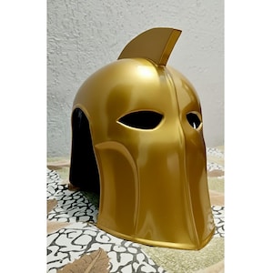 May include: A gold-coloured helmet with a distinctive fin on top and eye-shaped openings. The helmet has a smooth, glossy finish and a black interior. The design is reminiscent of a superhero or fantasy character.