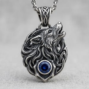 May include: A silver-colored pendant necklace featuring a howling wolf design. The wolf has intricate detailing and a blue gemstone at the base. The pendant is suspended from a silver chain, showcasing a Celtic knot design.