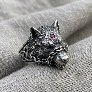May include: A silver ring featuring a detailed wolf head with a small red gemstone set on its forehead. The wolf is adorned with a delicate metal chain draped across its snout, and the sides of the ring band display an intricate knotwork pattern.