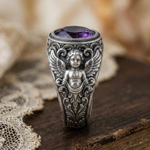 May include: A silver ring featuring a large, oval, purple gemstone. The ring is intricately detailed with an angel figure and ornate scrollwork. The ring's design is vintage-inspired, with a classic and elegant aesthetic.
