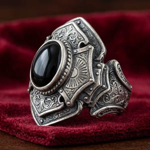 Puede incluir: Un anillo de plata detallado con una piedra negra ovalada. El anillo presenta intrincados patrones grabados y un diseño de armadura medieval. El anillo se exhibe sobre una superficie de terciopelo rojo, resaltando sus detalles y artesanía.