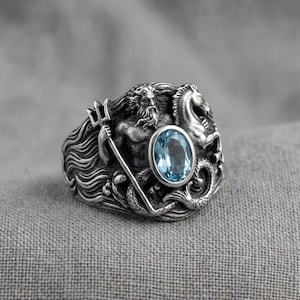 May include: A silver ring featuring a detailed relief of a bearded figure holding a trident next to a horse-like creature. The design includes flowing wave patterns and an oval-shaped, light blue gemstone set prominently in the center of the band.