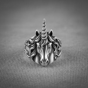 Könnte beinhalten: Ein silberfarbener Ring mit einem detaillierten Einhorn-Design. Der Ring zeigt den Kopf des Einhorns mit einem spiralförmigen Horn und fließender Mähne. Sterne sind um das Band herum eingraviert, was das Fantasy-Thema unterstreicht. Der Ring befindet sich vor einem grauen Hintergrund.