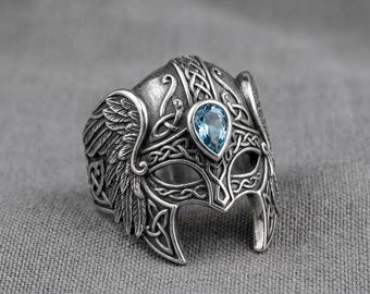 Anillo con forma de casco vikingo, piedra azul en forma de lágrima y diseño de nudo celta con alas.