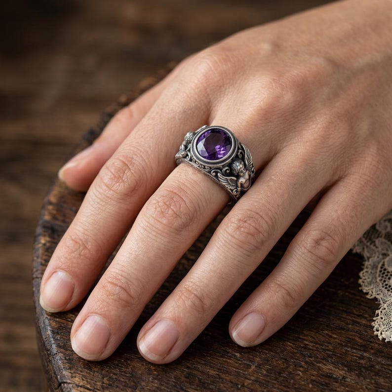 May include: A silver ring with a large, oval, purple gemstone. The ring has an ornate design with small figures and swirling patterns. The ring is on a person's finger, showcasing the intricate details and the gemstone's vibrant color.