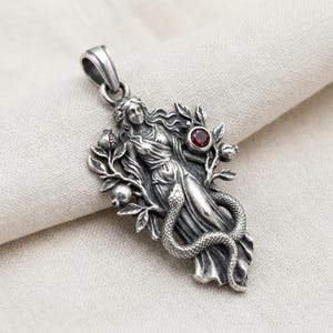 May include: A silver pendant featuring a sculpted figure with long flowing hair, surrounded by pomegranate branches and a coiled snake. A small, round, deep red gemstone is set into the metal near the figure's shoulder, with a bail at the top.
