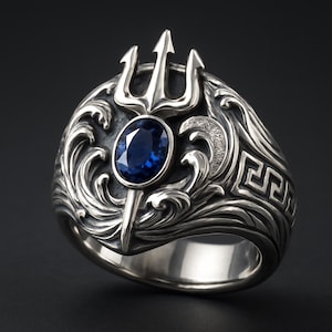 May include: A silver ring featuring a trident design with a blue oval gemstone at the center. The ring is intricately detailed with wave patterns and a Greek key border, set against a dark background.