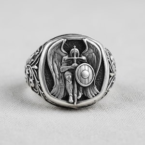 May include: A silver ring with a detailed relief of an angel warrior holding a sword and shield. The ring has an oval face with an ornate, floral design around the band. The angel has large wings and is in a defensive pose.