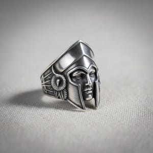 Könnte beinhalten: Ein silberner Ring mit einem detaillierten Design eines römischen Gladiatorenhelms. Der Ring hat ein markantes Gesicht auf der Vorderseite mit aufwendigen Details an Helm und Seiten. Der Ring besteht aus glänzendem Metall.