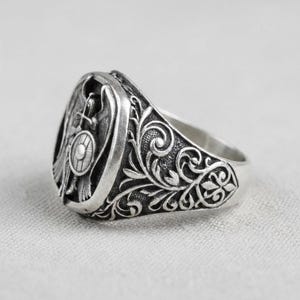 May include: A silver signet ring with an engraved design. The ring features a detailed emblem with a shield, cross, and figure, surrounded by ornate floral patterns. The ring is set against a neutral background.