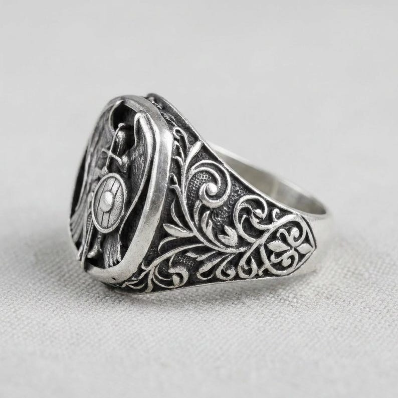 May include: A silver signet ring with an engraved design. The ring features a detailed emblem with a shield, cross, and figure, surrounded by ornate floral patterns. The ring is set against a neutral background.