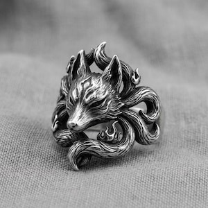 Può includere: Un anello color argento raffigurante una testa di volpe dettagliata con diverse code vorticose che avvolgono la fascia. Il metallo presenta una finitura ossidata per mettere in risalto le trame del pelo e i motivi a fiamma del design.