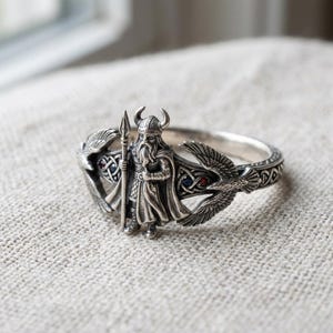 May include: A silver ring featuring a detailed Viking warrior with a spear, flanked by stylized eagle wings. The ring has intricate patterns and is set with small blue and red gemstones. The design is a detailed representation of Norse mythology.