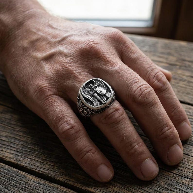 May include: A silver ring with a detailed design featuring a warrior with a sword and shield. The ring has an ornate, engraved band and is displayed on a hand.