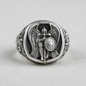 May include: A silver ring depicting an angel warrior with a sword and shield. The ring has an oval face with detailed relief and ornate side detailing. The dark background creates a striking contrast.