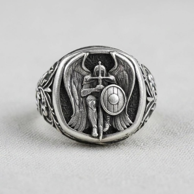 May include: A silver ring depicting an angel warrior with a sword and shield. The ring has an oval face with detailed relief and ornate side detailing. The dark background creates a striking contrast.
