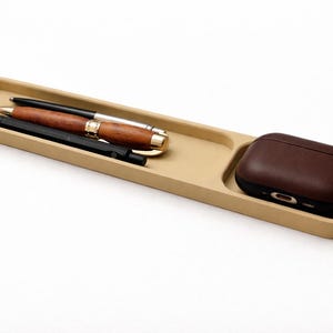 May include: A beige rectangular tray with rounded corners, holding pens and a brown case. The tray has two sections, one for pens and the other for the case. The pens include a wooden pen and two black pens. The case is dark brown.