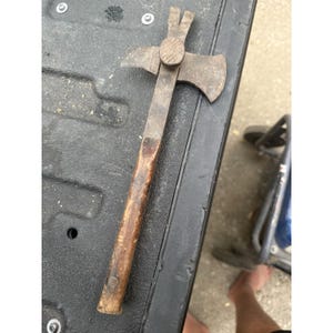 May include: A vintage multi-tool featuring a wooden handle and a metal head that combines an axe blade on one side, a hammer face in the center, and a nail puller claw at the top. The metal components show signs of oxidation and wear.