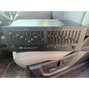 May include: A black Sansui SE-7 stereo graphic equalizer featuring two rows of vertical frequency sliders for left and right channels, a power button, tape monitor controls, and an output level knob on the front panel with printed text labels.