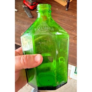 May include: A vintage, hexagonal, green glass bottle with the embossed word "Squibb". The bottle has a short neck and a flat, black base. The bottle is held in a hand.
