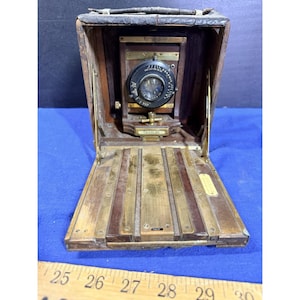 May include: A vintage folding camera constructed from dark wood and brass, featuring a black lens assembly labeled Graphic. The camera includes a leather-textured top handle and a metal base plate. A wooden ruler is visible, showing measurements in inches.