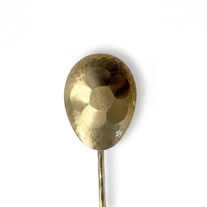 Könnte beinhalten: Ein goldfarbener, gehammerter Metalllöffel mit einem langen, schlanken Stiel. Die Löffelschale hat eine strukturierte, vertiefte Oberfläche, und auch der Stiel ist strukturiert. Der Löffel ist vor einem weißen Hintergrund platziert.