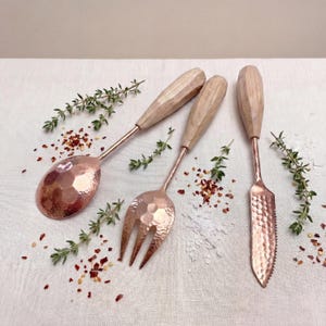 May include: A collection of four copper-toned serving utensils with wooden handles. The set includes a spoon, fork, and a knife with a serrated edge. The utensils are arranged on a white surface with herbs and spices.