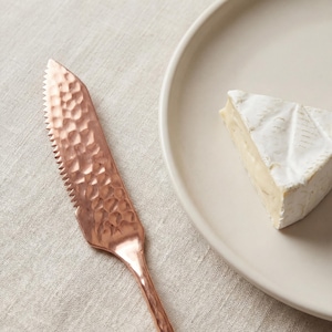Puede incluir: Un cuchillo de queso de cobre con un mango texturizado y un borde dentado, junto a una cuña de queso brie en un plato de color crema. El cuchillo tiene un diseño martillado y un mango largo y delgado. El queso es blanco cremoso con corteza.