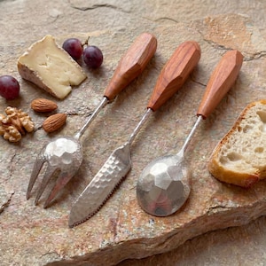 Könnte beinhalten: Ein Set aus drei Servierutensilien mit Holzgriffen und gehämmerten Metallköpfen. Das Set umfasst eine Gabel, ein Messer und einen Löffel. Die Utensilien sind auf einer Steinoberfläche mit Käse, Trauben, Nüssen und Brot angeordnet.