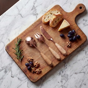 May include: A wooden serving board with a handle, featuring a selection of food items and copper-toned serving utensils. The board holds bread slices, cheese, grapes, walnuts, and rosemary. The utensils include a spoon, fork, and knife.