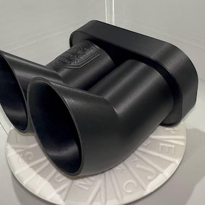 Peut inclure: Un embout d'échappement noir avec deux sorties inclinées. L'embout d'échappement est en matériau noir mat et comporte un emblème décoratif sur le dessus. L'embout d'échappement est posé sur une surface blanche.
