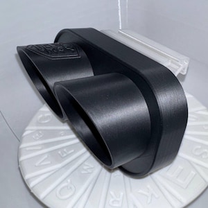 Pode incluir: Uma ponteira de escape preta de saída dupla com um design retangular. A ponteira de escape tem um acabamento brilhante e é montada numa base circular branca com letras. Uma peça de plástico transparente está presa no topo.