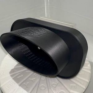 Puede incluir: Objeto negro curvado con interior texturizado y emblema en relieve. Tiene una base rectangular y una abertura ovalada redondeada. El emblema parece un escudo o logotipo. Probablemente es parte de un producto.