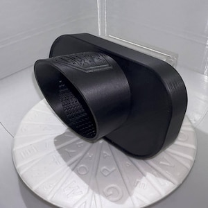 Könnte beinhalten: Ein schwarzes, 3D-gedrucktes Objekt mit einer zylindrischen Öffnung und einem rechteckigen Sockel. Der zylindrische Teil hat eine strukturierte Innenseite. Das Objekt liegt auf einer weißen, kreisförmigen Oberfläche mit geprägten Mustern. Der Hintergrund ist ein weißer, geschlossener Raum.