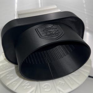 Op de afbeelding: Een zwart, cilindrisch object met een uitlopende opening, mogelijk een auto-onderdeel. Het object heeft de tekst "TCX WORKS" en een doodshoofdlogo in reliëf op de zijkant. Het staat op een witte, cirkelvormige basis met letters.