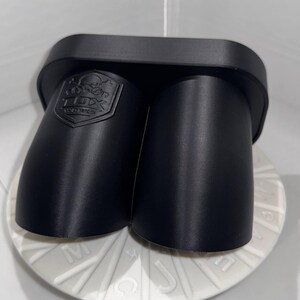 Peut inclure: Un embout d'échappement noir à double sortie avec une surface texturée. L'embout a un dessus rectangulaire et deux sorties cylindriques arrondies. Un logo est en relief sur le côté. Pour usage automobile.