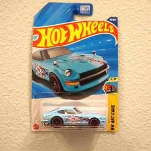 May include: A light blue Hot Wheels die-cast car with cherry blossom graphics. The car is packaged on a blue and yellow card with the Hot Wheels logo and "HW Art Cars" text. The car is marked as 4/10 and 62/250.