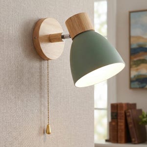 Puede incluir: Una lámpara de pared con base de madera y pantalla verde salvia. La lámpara tiene una cadena de tracción con un terminal dorado. La pantalla de la lámpara está inclinada hacia abajo y la luz está encendida.