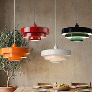 Può includere: Quattro moderne lampade a sospensione arancioni, rosse, bianche e nere con accenti verdi. Ogni lampada ha un design circolare a più livelli, sospeso dal soffitto. Le luci sono sopra un tavolo di legno con una fruttiera e delle posate.